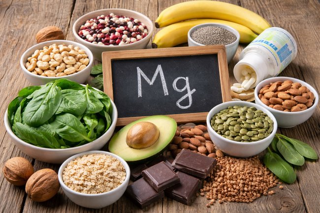 Magnesium-rich foods including spinach, pumpkin seeds, almonds, avocado, quinoa, dark chocolate, and nuts arranged around a chalkboard labeled Mg.