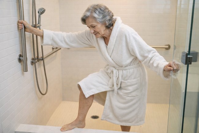 Senior woman in bathrobe stepping into shower while holding grab bar for safety